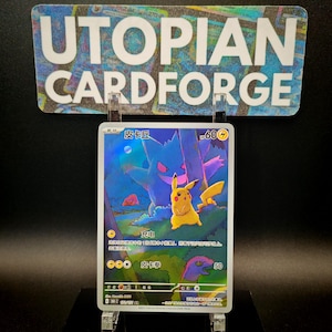 May include: A holographic Pokémon trading card featuring Pikachu and Gengar in a forest scene. The card has Japanese text and a 60-point value. The card is displayed in a clear acrylic holder, with a sign above that reads "UTOPIAN CARDFORGE."
