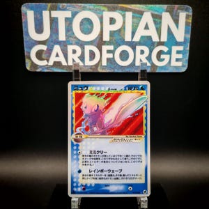 May include: A holographic Pokémon trading card featuring the character Mew. The card has a blue border and Japanese text. The card is displayed on a clear stand in front of a sign that reads "UTOPIAN CARDFORGE".