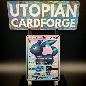 May include: A Pokémon trading card featuring Umbreon GX. The card has a black and blue Umbreon illustration with the text "Umbreon GX" and "HP 200". The card is displayed in a clear holder with the text "UTOPIAN CARDFORGE" in the background.