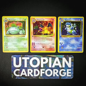 Puede incluir: Tres cartas coleccionables de Pokémon con Venusaur, Charizard y Blastoise. Las cartas tienen bordes amarillos e ilustraciones coloridas de los Pokémon. En la parte inferior hay una pegatina rectangular con el texto "UTOPIAN CARDFORGE".