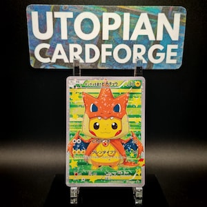 May include: A Pokémon trading card featuring Pikachu in an orange costume with a flame-like design. The card has Japanese text and is displayed on a clear stand. The text "Utopian Cardforge" is above the card.