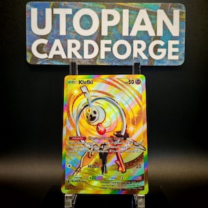 Może przedstawiać: Holograficzna karta Pokémon Klefki ze złotym i żółtym tłem. Karta przedstawia wzór w kształcie klucza z czerwonym akcentem oraz tekst "Klefki" i "Hook". Karta jest wyświetlana na przezroczystym stojaku z napisem "UTOPIAN CARDFORGE" nad nią.