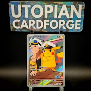 May include: A holographic trading card featuring a cartoon image of a man and a yellow Pikachu wearing a hat. The card has Japanese text and is displayed in a clear acrylic holder. The sign above the card reads "UTOPIAN CARDFORGE."