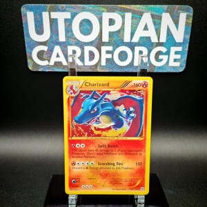 May include: A Charizard Pokémon trading card with a yellow border. The card features a blue and red dragon-like creature. The card has the text "Charizard" and "HP 160". The card is displayed in a clear holder with the text "UTOPIAN CARDFORGE" in the background.