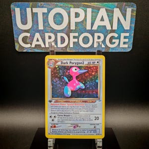 Könnte beinhalten: Eine Dark Porygon2 Pokémon-Sammelkarte mit gelbem Rand. Die Karte zeigt eine rosa und blaue Cartoonfigur. Die Karte wird in einem transparenten Kunststoffhalter vor einem schwarzen Hintergrund präsentiert. Der Text "UTOPIAN CARDFORGE" ist oben sichtbar.