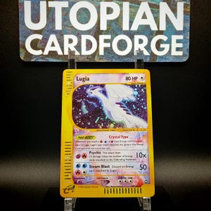 Peut inclure: Une carte à collectionner Pokémon Lugia jaune avec une illustration blanche et bleue de Lugia. La carte porte les mentions "Lugia", "80 HP" et "Psychic". La carte est présentée sur un support transparent devant une enseigne "UTOPIAN CARDFORGE".