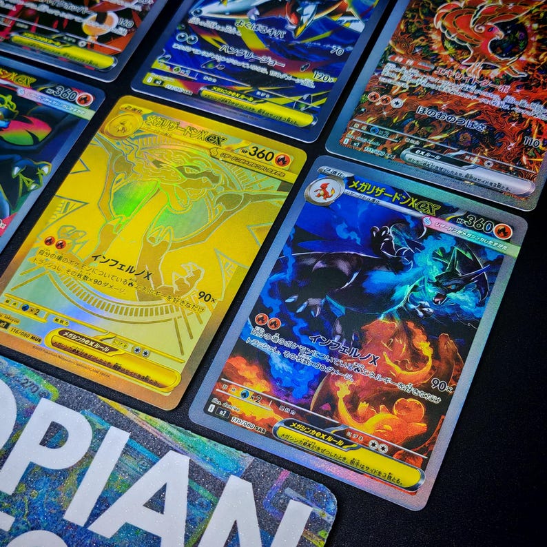 May include: A collection of holographic trading cards featuring various characters and designs. The cards have a shiny, reflective surface with Japanese text and vibrant colors, including gold, blue, and red. The cards are displayed on a dark surface.