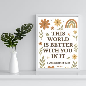 May include: A framed print with the text "THIS WORLD IS BETTER WITH YOU IN IT" and the Bible verse 1 Corinthians 12:18. The design includes floral and rainbow illustrations in shades of brown, orange, and green. A white vase with green leaves is on the left.