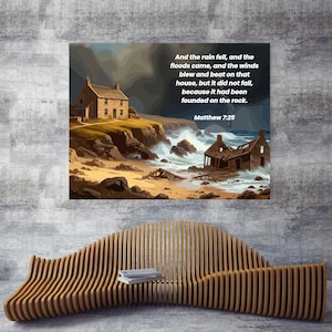 Puede incluir: Una impresión enmarcada que representa una escena costera con una casa en un acantilado, olas rompiendo y texto de Mateo 7:25. La obra de arte se exhibe sobre un banco de madera con una pila de libros.