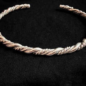 May include: A circular open cuff bracelet featuring a twisted design with alternating strands of polished silver-toned and copper-toned metal. The bracelet has a continuous braided texture throughout its entire length, creating a metallic rope effect.