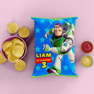 Puede incluir: Una bolsa de patatas fritas azul con Buzz Lightyear de Toy Story, con el texto "LIAM IS TURNING 3". La bolsa est&aacute; rodeada de patatas fritas en un cuenco rosa, patatas fritas sueltas y peque&ntilde;os cuencos con salsas rojas y amarillas. El fondo es rosa liso.