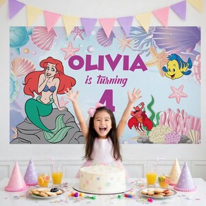 May include: A rectangular party backdrop featuring a light blue underwater scene with a mermaid, a yellow fish, a red crab, seashells, and starfish. The text reads OLIVIA is turning 4 in purple letters. A colorful triangular bunting hangs above.