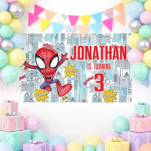 May include: A birthday banner with a Spider-Man graphic, the name "Jonathan", and the number "3". The banner is set against a cityscape backdrop and decorated with balloons and wrapped presents. The text on the banner reads "Jonathan is turning 3".
