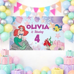May include: A rectangular party backdrop featuring a cartoon mermaid with red hair, a yellow fish, and a red crab. The text reads OLIVIA is turning 4. The background displays an underwater scene with seashells, starfish, and coral, framed by colorful balloons and a triangular bunting banner.