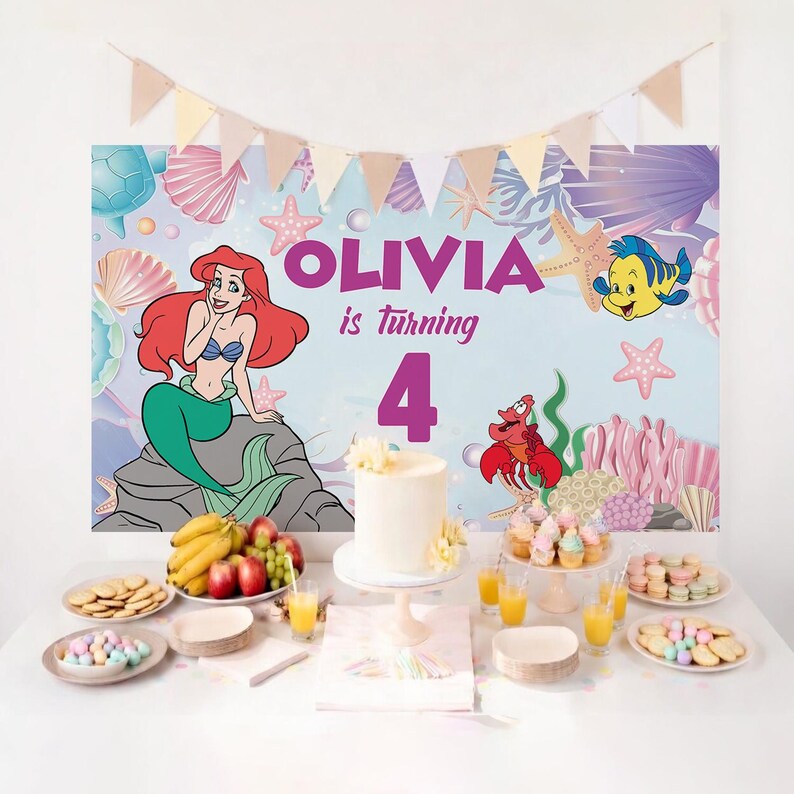 May include: A party backdrop featuring Ariel the mermaid, Flounder, and Sebastian on a light blue background with the text OLIVIA is turning 4. The scene includes a white cake, fruit, cupcakes, and cookies on a table with a triangular banner.