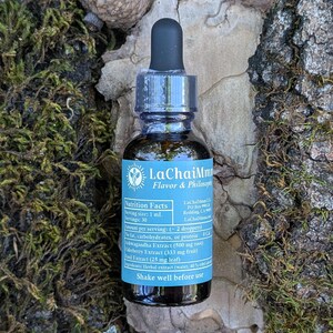May include: A small, dark glass bottle with a black dropper top, labeled "LaChaiMm Flavor & Philosophy." The label includes nutrition facts and ingredient information. The bottle is surrounded by moss, leaves, and an acorn.