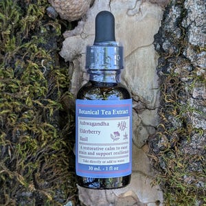 May include: A dark glass bottle of botanical tea extract with a black dropper. The label reads "Ashwagandha Elderberry Basil" and "30 mL - 1 fl oz." The bottle is on a bed of moss and leaves, with an acorn in the background.