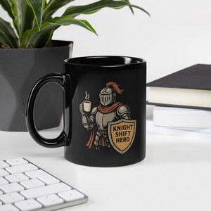 May include: Black ceramic mug featuring a knight in armor holding a cup of coffee. The knight is depicted with a red plume and scarf, and a shield that reads "KNIGHT SHIFT HERO". The mug has a large black handle.