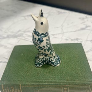 May include: A white ceramic bird figurine with green leaf and floral patterns. The bird has an open beak and is on a green book. The figurine is approximately 10 cm tall. Decorative ceramic bird ornament.