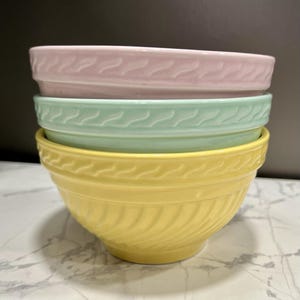 Könnte beinhalten: Ein Stapel von drei Keramik-Rührschüsseln in Pastellfarben: Rosa, Mintgrün und Gelb. Jede Schüssel hat eine strukturierte, geriffelte Außenseite und ein dekoratives Muster am Rand. Die Schüsseln sind ineinander verschachtelt.