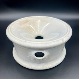 May include: A light gray ceramic oil diffuser with a wide, shallow bowl and a central hole. The diffuser has a rounded base and a smaller hole on the side. The ceramic has a speckled, textured appearance.