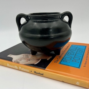 Könnte beinhalten: Ein kleiner, schwarzer Keramiktopf mit zwei Henkeln und drei Füßen. Der Topf steht auf einem Buch mit dem Titel "Roseville Pottery". Das Buch hat einen orangefarbenen Einband mit blauem und weißem Text.