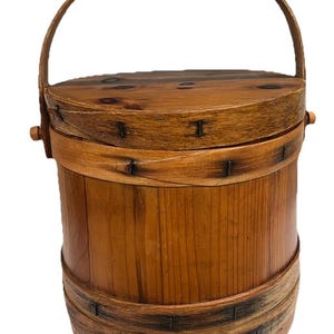 May include: A cylindrical wooden bucket with a matching lid and a curved handle. The container features visible wood grain, dark knots on the lid, and thin wooden bands secured with small metal nails around the body and the lid's edge.