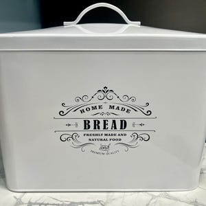 Op de afbeelding: Een witte rechthoekige broodtrommel met een gebogen handvat op het deksel. De voorkant van de trommel heeft zwarte tekst met de tekst "HOME MADE BREAD" en "FRESHLY MADE AND NATURAL FOOD and PREMIUM QUALITY".