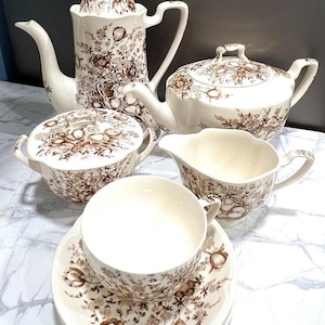 May include: A vintage tea set with a floral pattern in shades of brown and cream. The set includes a teapot, a creamer, a sugar bowl, and a cup and saucer set. The tea set is made of ceramic.