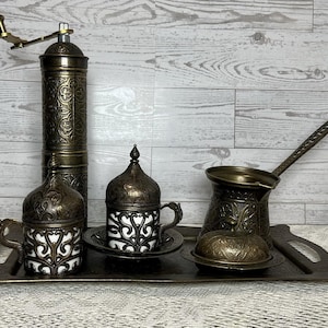 May include: A brass coffee set featuring a tall cylindrical grinder with a hand crank, two cups with lids and saucers, a long-handled pot, and a small lidded bowl, all arranged on a rectangular tray, featuring intricate embossed metal patterns.