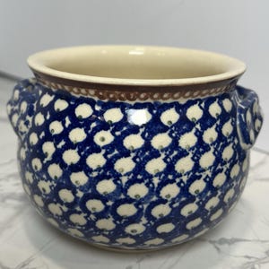May include: A round ceramic bowl with two side handles, featuring a cream-colored base decorated with a repeating pattern of dark blue teardrop shapes, each containing a small green dot. The rim is accented with a brown band and a dotted border.
