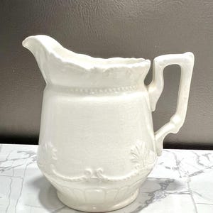 May include: A white ceramic pitcher with a handle and spout. The pitcher has a decorative floral design around the base and a beaded trim near the top. The pitcher is likely used for serving beverages.