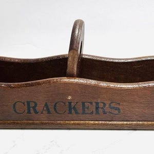 May include: A long, rectangular dark brown wooden server with a central handle and wavy side edges. The word CRACKERS is printed in black capital letters on the front panel. The server is divided into two equal compartments by the central handle.