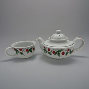 Peut inclure: Une théière en porcelaine blanche avec une tasse assortie, toutes deux ornées d'un motif répétitif de fraises rouges et de feuilles vertes sur de fines tiges brunes. La théière présente un couvercle arrondi avec un bouton et une anse courbée.