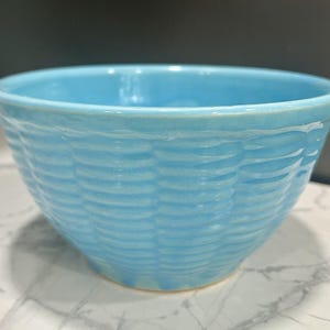 Puede incluir: Un cuenco de cerámica azul claro con un diseño de tejido de cesta. El cuenco tiene forma redondeada y un acabado liso y brillante. El cuenco está sobre una superficie de mármol blanco y gris.