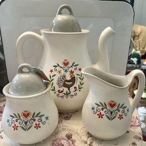 Op de afbeelding: Een crèmekleurige keramische theeset met een gespikkelde afwerking. De set bevat een theepot, een melkkan en een suikerpot, elk versierd met een kleurrijk bloemen- en hartontwerp en een hanenillustratie. De theepot heeft een gebogen tuit en handvat.