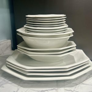 May include: A stack of white ceramic dinnerware, including plates and bowls. The plates have a unique octagonal shape with a beaded edge. The bowls are round with a similar octagonal shape. The set is arranged on a marble countertop.
