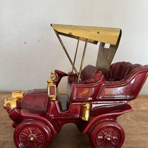 Peut inclure: Figurine de voiture ancienne en céramique bordeaux avec des accents dorés. La voiture présente un design détaillé, comprenant des roues, un siège conducteur et un auvent doré. La figurine est exposée sur une surface en bois.