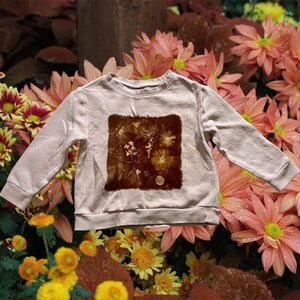 Puede incluir: Una sudadera beige claro con cuello redondo y mangas largas. Presenta un gráfico cuadrado marrón de flores y follaje prensados. El fondo está lleno de flores coloridas en tonos rosa, amarillo y rojo.