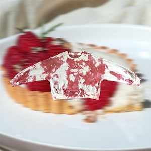 May include: A white sweatshirt with a red tie-dye pattern. The long-sleeved top has a crew neck and ribbed cuffs and hem. The shirt is displayed over a blurred image of a dessert.