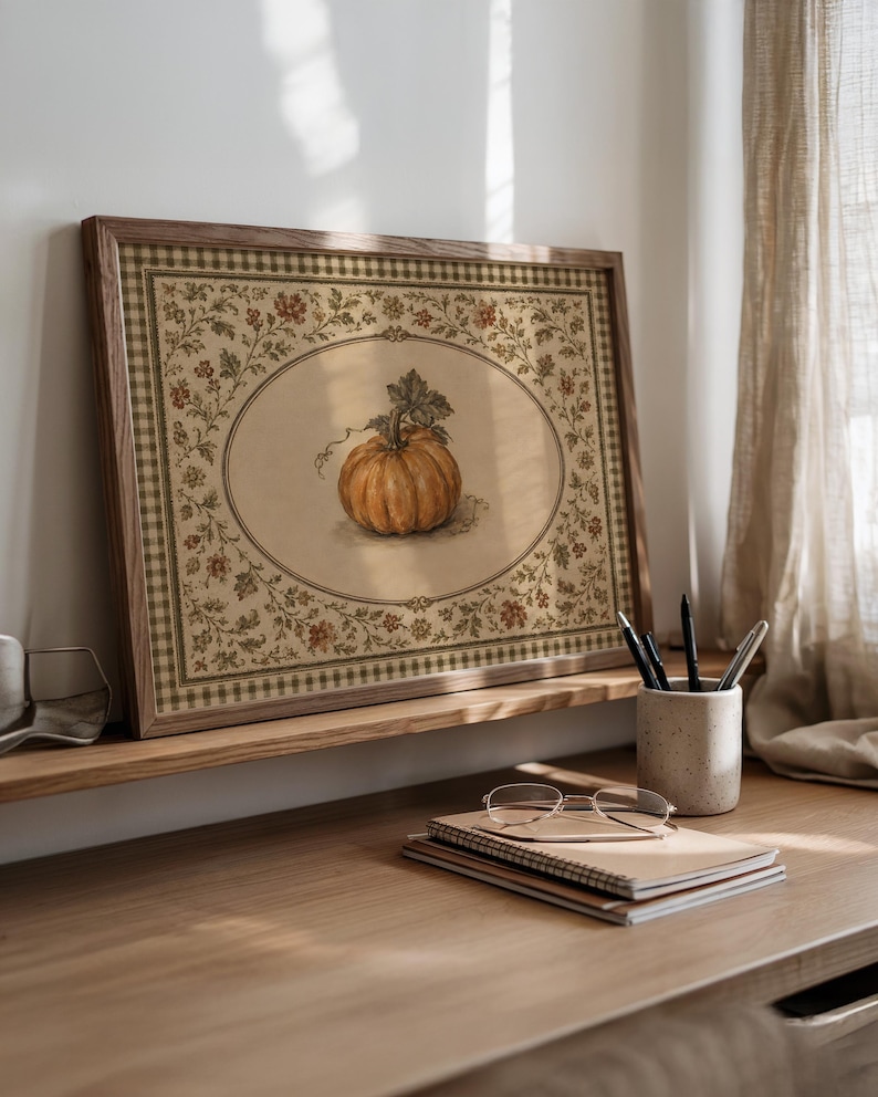 May include: A rectangular framed print featuring a central illustration of an orange pumpkin inside an oval border. The surrounding area displays a detailed floral pattern with green leaves and small red flowers, finished with a green gingham border.