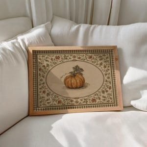 May include: A rectangular wooden frame containing a print of a single orange pumpkin centered within an oval border. The print features a detailed floral vine pattern surrounding the oval and a checkered border edge in muted earth tones.