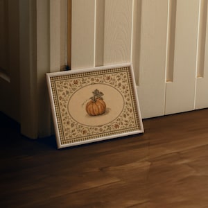 May include: A rectangular framed print featuring a central illustration of an orange pumpkin with green leaves. The artwork is surrounded by a decorative border of small floral and leaf patterns in muted earth tones, enclosed in a white frame.