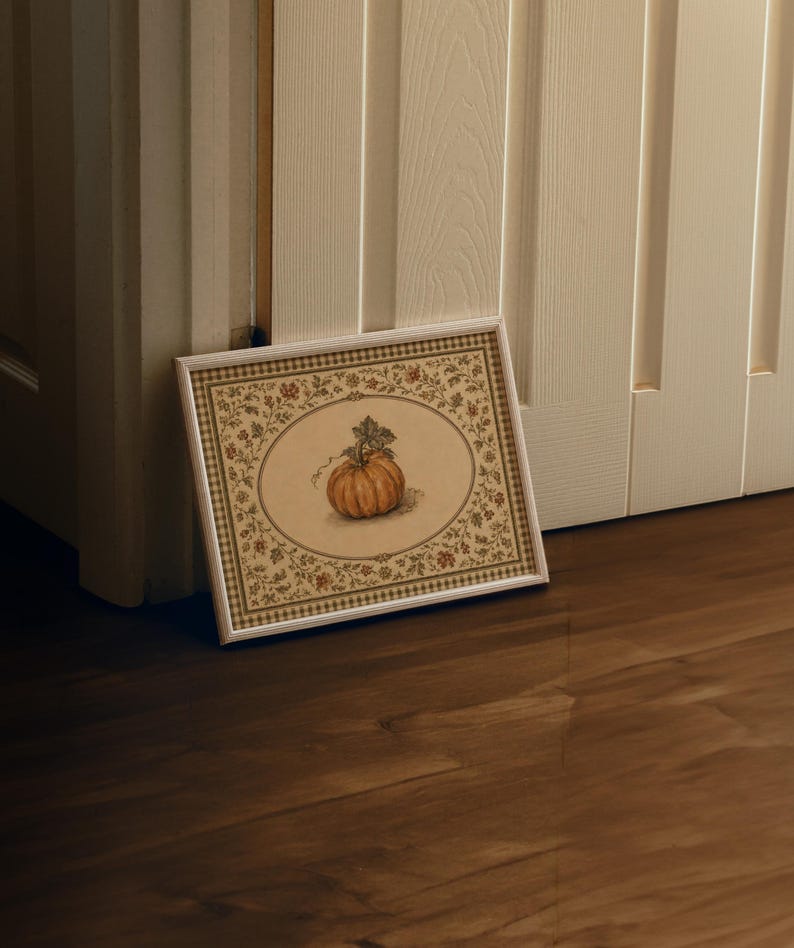 May include: A rectangular framed print featuring a central illustration of an orange pumpkin with green leaves. The artwork is surrounded by a decorative border of small floral and leaf patterns in muted earth tones, enclosed in a white frame.