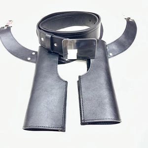 May include: Black leather belt with a silver buckle and two attached leather pieces. The belt is rolled up and the leather pieces are curved. The belt and leather pieces are on a white background.