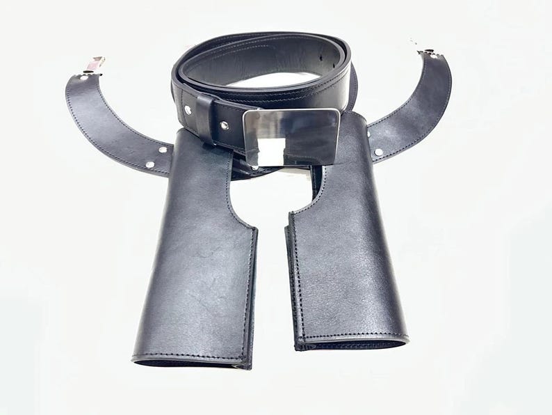 May include: Black leather belt with a silver buckle and two attached leather pieces. The belt is rolled up and the leather pieces are curved. The belt and leather pieces are on a white background.