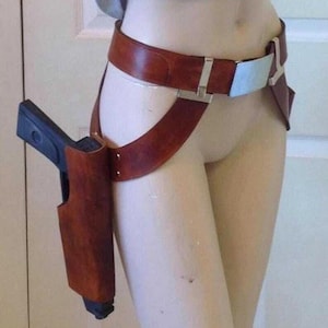May include: A brown leather holster with a black handgun, attached to a brown leather belt and thigh strap. The belt has a silver buckle and a light blue accent. The holster is designed to be worn on the thigh.