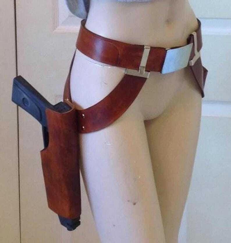 May include: A brown leather holster with a black handgun, attached to a brown leather belt and thigh strap. The belt has a silver buckle and a light blue accent. The holster is designed to be worn on the thigh.