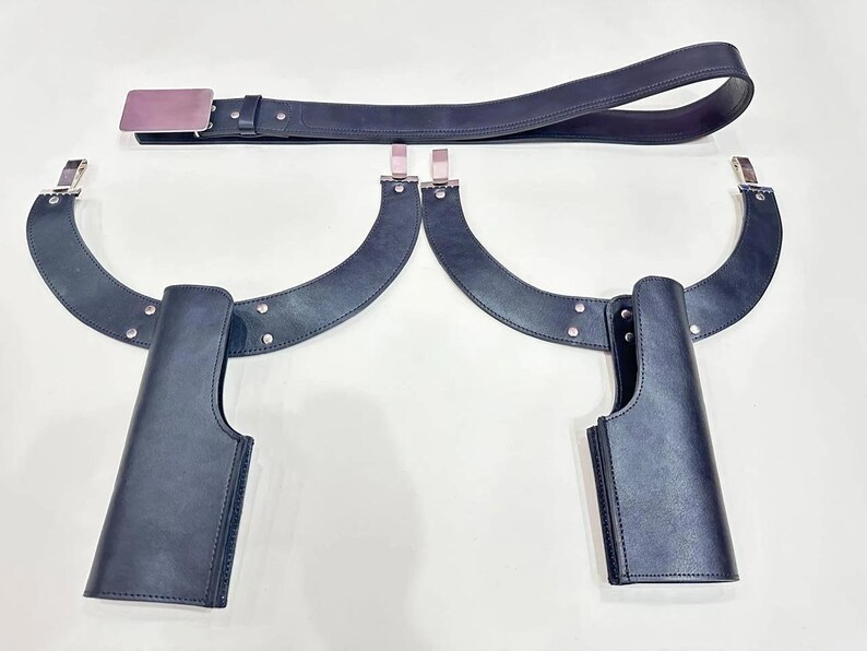 May include: A black leather belt with a silver buckle and two matching black leather holsters. The holsters are attached to curved black leather supports with silver hardware. The items are displayed on a white surface.