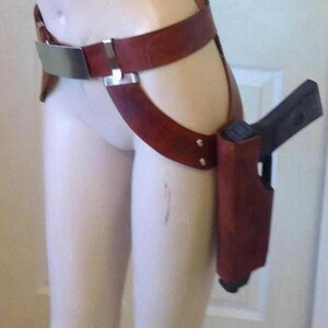 May include: A brown leather thigh holster with a gun. The holster is attached to a belt with a silver buckle and a strap that goes around the thigh. The gun is black.